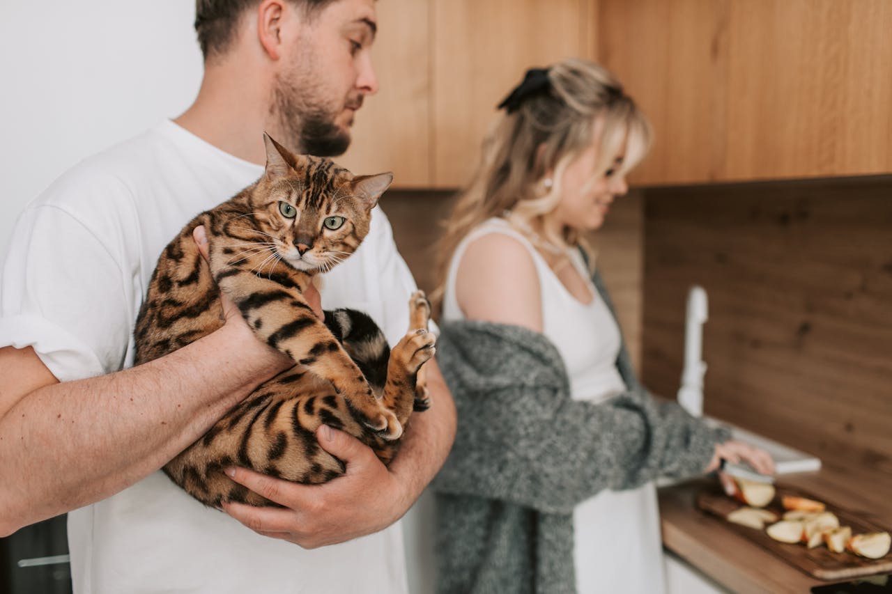 A man holding a Bengal cat while a woman slices fruits in a cozy kitchen setting.